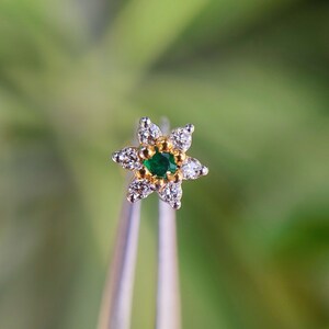 May include: A gold flower-shaped nose ring with a green gemstone in the center and clear diamonds around the edges.