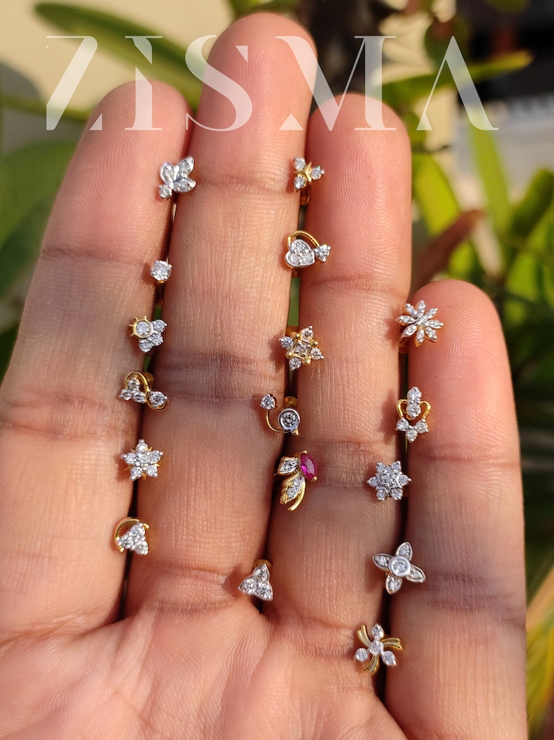 May include: A collection of 14 gold nose rings with various designs featuring small diamonds. The designs include a heart, a flower, a star, a leaf, and a swirl.