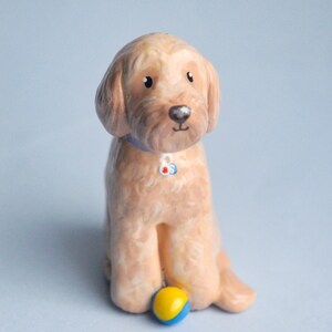 May include: A ceramic figurine of a golden-colored dog with a blue collar and a small ball. The dog is sitting with its front paws forward and a yellow and blue ball in front of it. The background is a light blue.