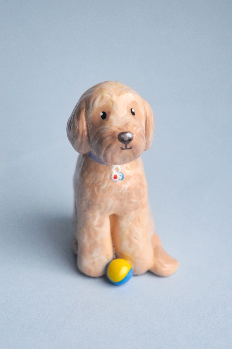 May include: A ceramic figurine of a golden-colored dog with a blue collar and a small ball. The dog is sitting with its front paws forward and a yellow and blue ball in front of it. The background is a light blue.