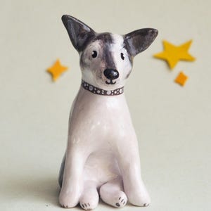 May include: A ceramic dog figurine with a white body, gray and black ears, and a brown dotted collar. The dog is sitting with its legs tucked in. Two yellow star-shaped decorations are in the background.
