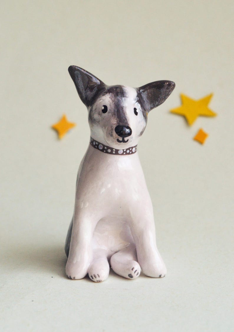 May include: A ceramic dog figurine with a white body, gray and black ears, and a brown dotted collar. The dog is sitting with its legs tucked in. Two yellow star-shaped decorations are in the background.
