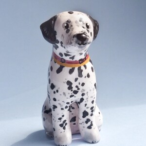 May include: A ceramic Dalmatian dog figurine with black spots on a white body. The dog is sitting with a red and yellow collar. The ears and the area around the eyes are black. The figurine is on a light blue background.