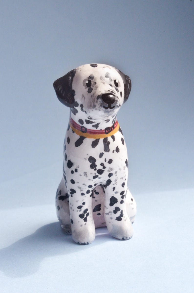 May include: A ceramic Dalmatian dog figurine with black spots on a white body. The dog is sitting with a red and yellow collar. The ears and the area around the eyes are black. The figurine is on a light blue background.