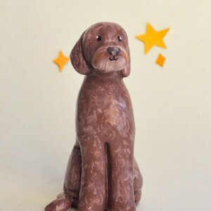 May include: A brown ceramic dog figurine in a seated position, with a light brown muzzle and floppy ears. The dog has a textured coat and is set against a cream background with yellow star accents. A charming decorative item.