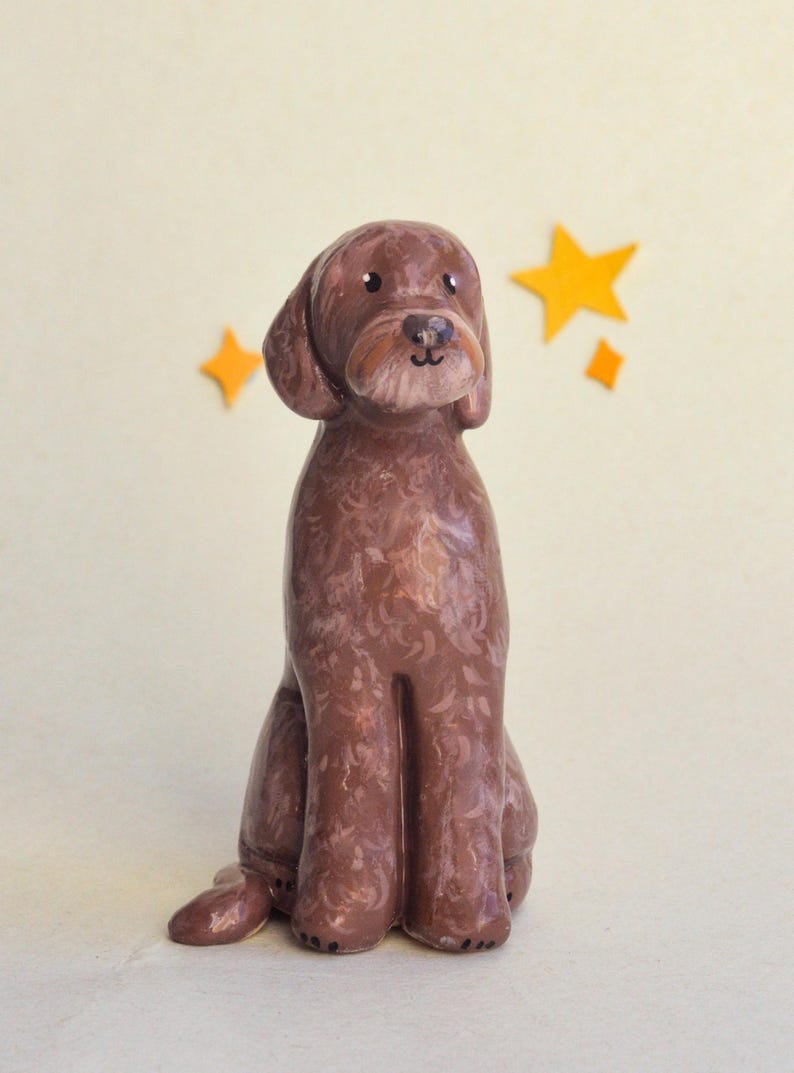May include: A brown ceramic dog figurine in a seated position, with a light brown muzzle and floppy ears. The dog has a textured coat and is set against a cream background with yellow star accents. A charming decorative item.