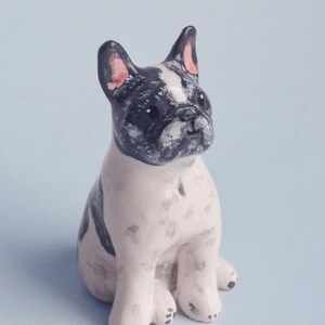 May include: A ceramic figurine of a French Bulldog in a sitting position. The dog has a white body with black markings on its head and back. The ears are pink-lined. The figurine is on a light blue background.