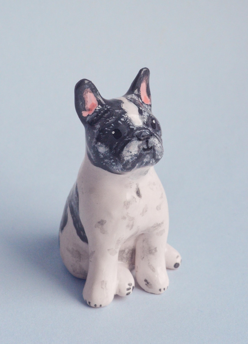 May include: A ceramic figurine of a French Bulldog in a sitting position. The dog has a white body with black markings on its head and back. The ears are pink-lined. The figurine is on a light blue background.