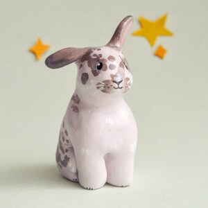 May include: A ceramic bunny figurine in a seated pose, with a pink body and brown spotted markings. The bunny has brown ears and is set against a light green background with yellow star accents. A whimsical decorative item.