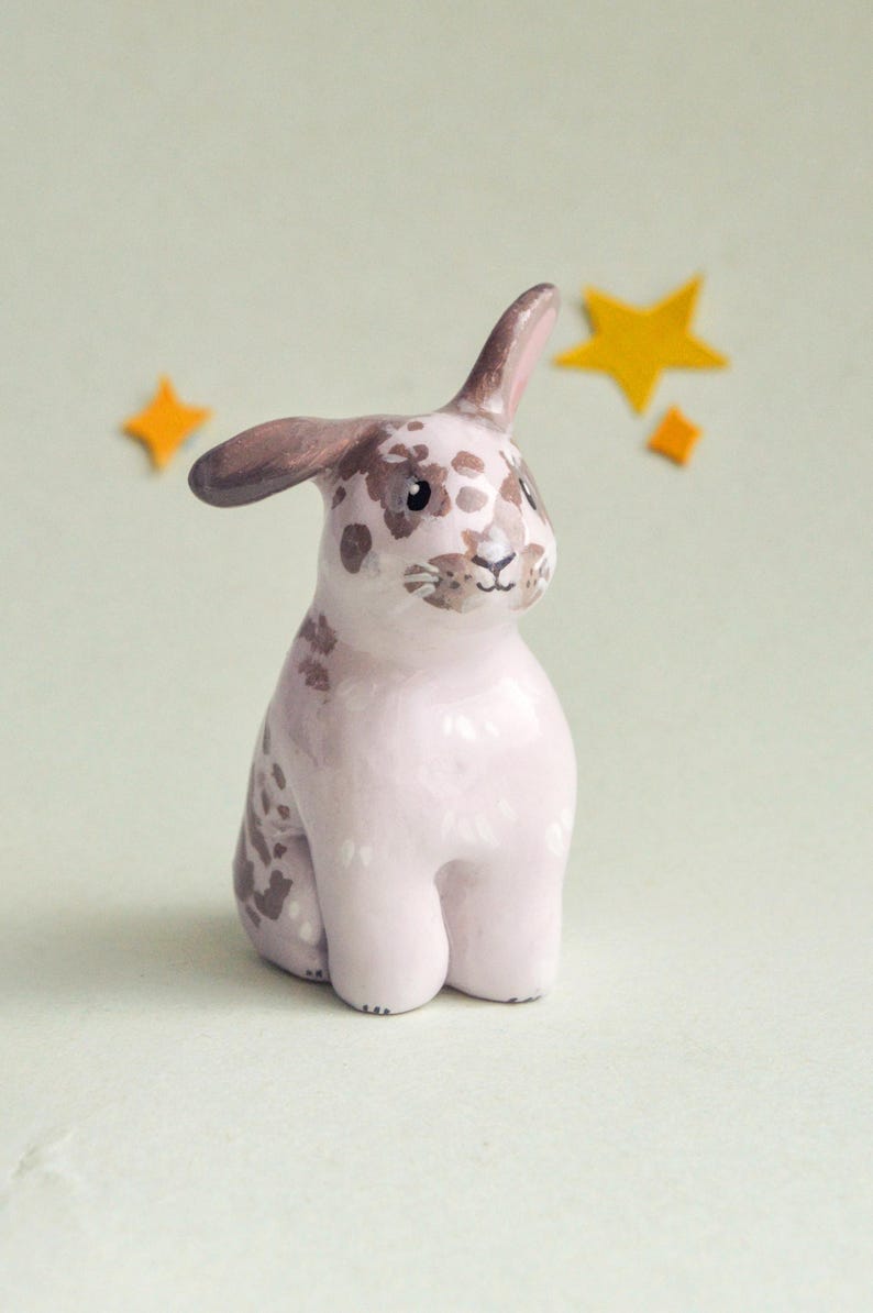 May include: A ceramic bunny figurine in a seated pose, with a pink body and brown spotted markings. The bunny has brown ears and is set against a light green background with yellow star accents. A whimsical decorative item.