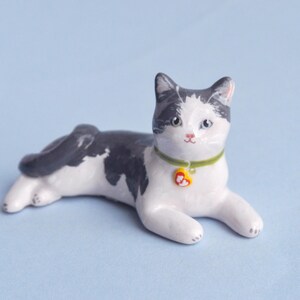 May include: A ceramic cat figurine in a relaxed pose, featuring a white body with black markings on its head and back. It wears a green collar with a yellow heart-shaped tag. The cat is on a light blue surface.