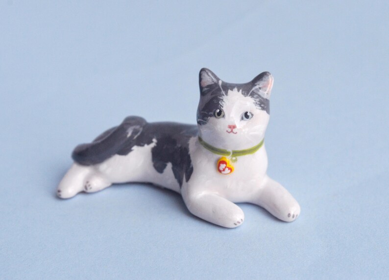 May include: A ceramic cat figurine in a relaxed pose, featuring a white body with black markings on its head and back. It wears a green collar with a yellow heart-shaped tag. The cat is on a light blue surface.