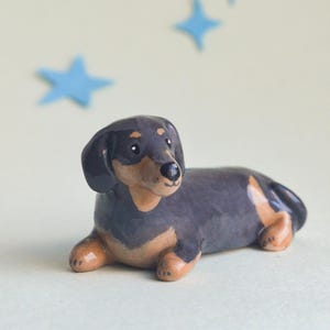May include: A ceramic dachshund figurine in a resting pose. The dog is primarily black with tan markings on its legs and belly. Blue star-shaped decorations are in the background.