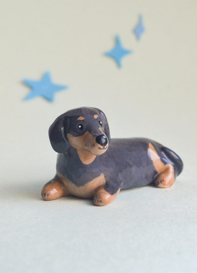 May include: A ceramic dachshund figurine in a resting pose. The dog is primarily black with tan markings on its legs and belly. Blue star-shaped decorations are in the background.