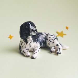 May include: A ceramic figurine of a dog, lying down with its front paws extended. The dog has a black head and ears, with a white body speckled with black spots. The background is a light green color, with three yellow star-shaped decorations.