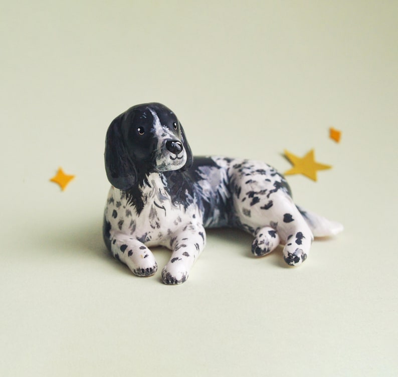May include: A ceramic figurine of a dog, lying down with its front paws extended. The dog has a black head and ears, with a white body speckled with black spots. The background is a light green color, with three yellow star-shaped decorations.
