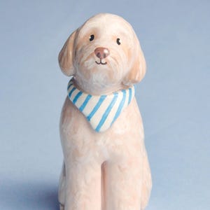 May include: A ceramic dog figurine in a seated position, with a light tan coat and a blue and white striped bandana. The dog has a pink nose and dark eyes, set against a light blue background.
