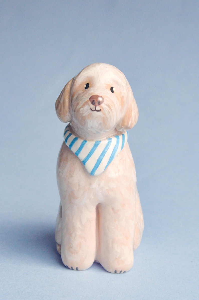May include: A ceramic dog figurine in a seated position, with a light tan coat and a blue and white striped bandana. The dog has a pink nose and dark eyes, set against a light blue background.