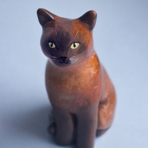 May include: A ceramic cat figurine in shades of brown and dark brown, with yellow eyes and white whiskers. The cat is sitting upright, with its ears perked up, and is set against a light blue background.