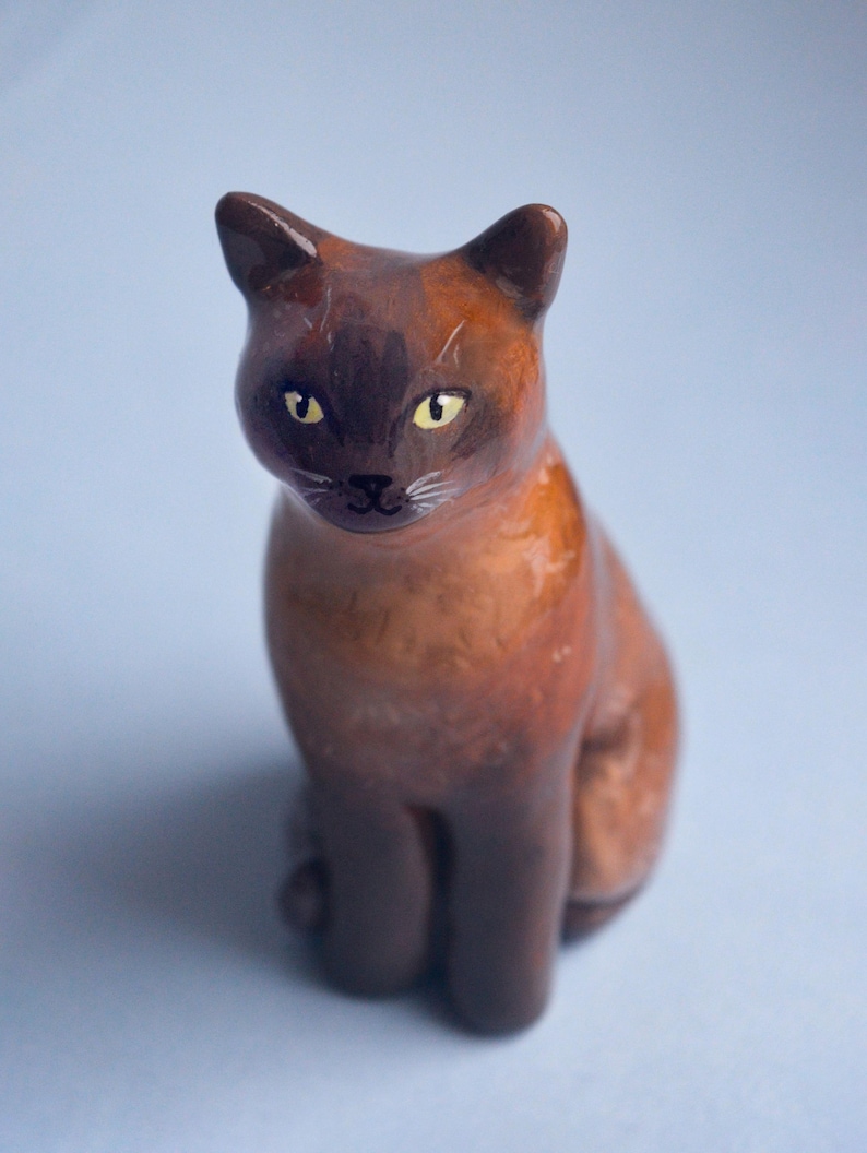 May include: A ceramic cat figurine in shades of brown and dark brown, with yellow eyes and white whiskers. The cat is sitting upright, with its ears perked up, and is set against a light blue background.