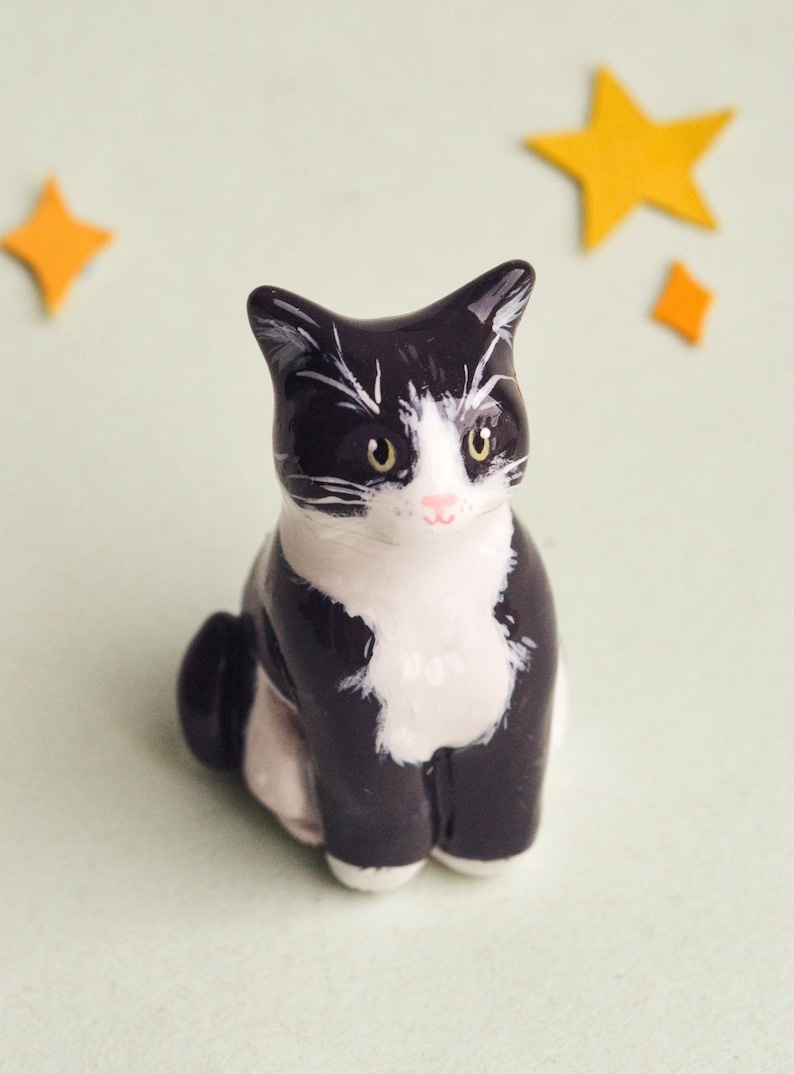 May include: A ceramic figurine of a sitting cat with black and white markings. The cat has yellow eyes and a pink nose. Two yellow star-shaped decorations are in the background. The figurine is on a light green surface.