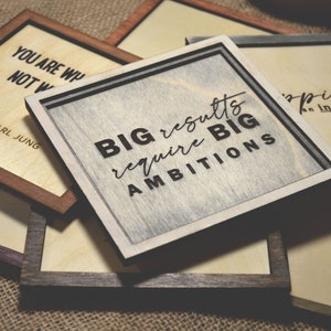 May include: Four wooden signs with inspirational quotes. The top sign is brown with the quote "You are what you do, not what you say." by Carl Jung. The second sign is gray with the quote "Big results require big ambitions." The third sign is brown with the quote "Happiness is an inside job." The fourth sign is brown with a blank surface.