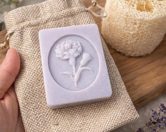 May include: A lavender soap bar with a carved flower design, on a beige linen bag. A natural loofah sponge and lavender sprig are also present. The soap is rectangular with a floral relief. The items are arranged on a wooden board.
