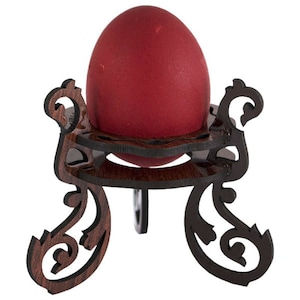 May include: A red egg sits in a wooden egg holder with a decorative, ornate design. The holder has three legs and a circular top.
