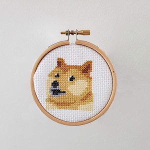 May include: A cross-stitch embroidery of the Doge meme, a Shiba Inu dog with a surprised expression, framed in a round wooden hoop.