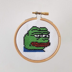 May include: A green frog meme cross-stitch in a wooden embroidery hoop. The frog has a green body, black eyes, and a red mouth.