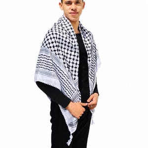 May include: A black and white keffiyeh scarf with a geometric pattern. The scarf is draped around the shoulders. The scarf has a fringe at the ends. The person in the image is wearing a black long-sleeve shirt and black pants.