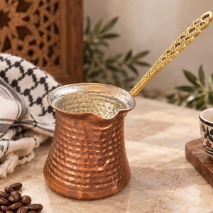 May include: A hammered copper coffee pot with a wide base, a narrow neck, and a pouring spout. The pot features a long, ornate, gold-toned metal handle with an intricate cutout pattern. The interior of the pot has a silver-colored metallic finish.