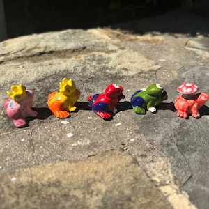 May include: Five small ceramic frog figurines in various colors, including yellow, red, blue, green, and pink. Each frog has a unique design and is decorated with white polka dots.