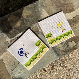May include: Two white ceramic tiles with a pixelated design of a video game character. The character on the left tile is wearing a blue hat and the character on the right tile is wearing a yellow hat. Both characters are jumping over a green platform.