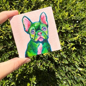 May include: A small square painting of a green French Bulldog with pink accents on a light pink background. The dog's eyes are blue, and its ears and nose are pink. The artwork is held against a backdrop of green foliage.