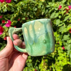 May include: A green ceramic mug with a unique, drippy glaze pattern. The mug has a handle and is perfect for enjoying a warm beverage.