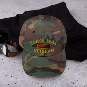 May include: A camouflage baseball cap with the text "Class War Veteran" in yellow embroidery and a red, black, and yellow flag design.