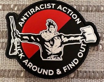Può includere: Un cerotto circolare nero e rosso con il testo "Antiracist Action" e "Stick Around & Find Out" in bianco. Una silhouette bianca di una persona che tiene un fucile e un libro è al centro.