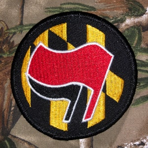 May include: A black and gold embroidered patch with a red fist raised in the air. The fist is outlined in white and is set against a background of black and gold stripes.