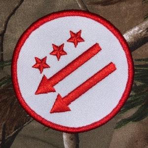 Op de afbeelding: Een witte cirkelvormige patch met een rode rand. Binnen de cirkel bevinden zich drie rode vijfpuntige sterren en twee rode pijlen die naar rechts wijzen.