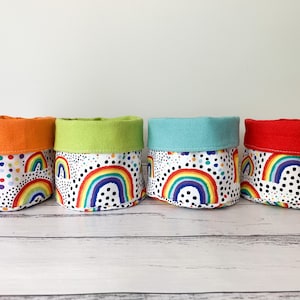 May include: Four fabric storage containers with a rainbow and polka dot design. Each container has a different coloured top: orange, green, blue, and red. The containers are cylindrical and sit on a white wooden surface.