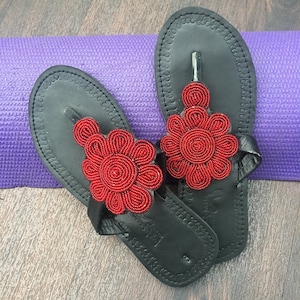 May include: A pair of black leather sandals with red beaded flower embellishments. The sandals have a simple thong strap design and are perfect for summer.