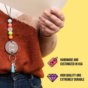 May include: A wooden keychain with the text "Mrs. King" on it. The keychain is attached to a blue and white beaded lanyard with a blue tassel. The lanyard is worn by a person wearing a brown sweater and blue jeans. The image also includes two icons: one with a hand holding a heart and the text "Handmade and Customized in USA" and the other with a diamond and the text "High Quality and Extremely Durable."