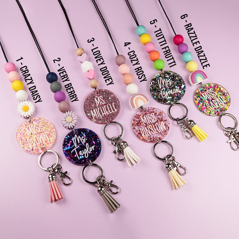 May include: Ten colourful lanyards with different bead patterns and personalised name tags. Each lanyard features a charm with a rainbow, a flower, or glitter. The name tags have a variety of colours and glitter accents.