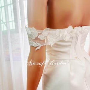 May include: White off-the-shoulder dress with delicate floral lace detailing on the sleeves. The dress has a fitted bodice and a flowing skirt.