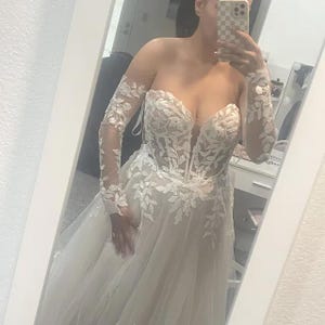 May include: A strapless ivory wedding gown with a sweetheart neckline and a flowing skirt. The bodice is adorned with intricate floral lace, and the long sleeves are sheer, also featuring lace detailing. The dress is captured in a mirror reflection.