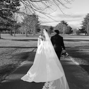 May include: Black and white photograph of a wedding. A person in a white wedding dress with a long veil walks down a path, followed by another person in a black suit. The veil has a lace trim. The background shows trees and a grassy area.