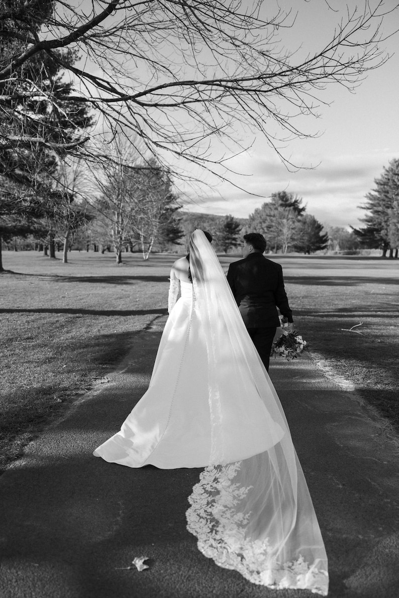 May include: Black and white photograph of a wedding. A person in a white wedding dress with a long veil walks down a path, followed by another person in a black suit. The veil has a lace trim. The background shows trees and a grassy area.