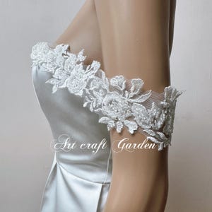 Könnte beinhalten: Ein weißes, schulterfreies Kleid mit zarten Spitzendetails am Oberteil und an den Ärmeln. Die Spitze ist ein Blumenmuster auf weißem Hintergrund. Das Kleid ist aus einem glatten, satinartigen Stoff gefertigt.