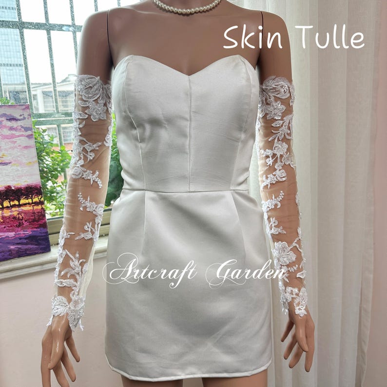 May include: A white satin mini dress with a sweetheart neckline and a fitted bodice. The dress is paired with sheer, long sleeves adorned with white floral lace. The text "Skin Tulle" and "Artcraft Garden" are visible.